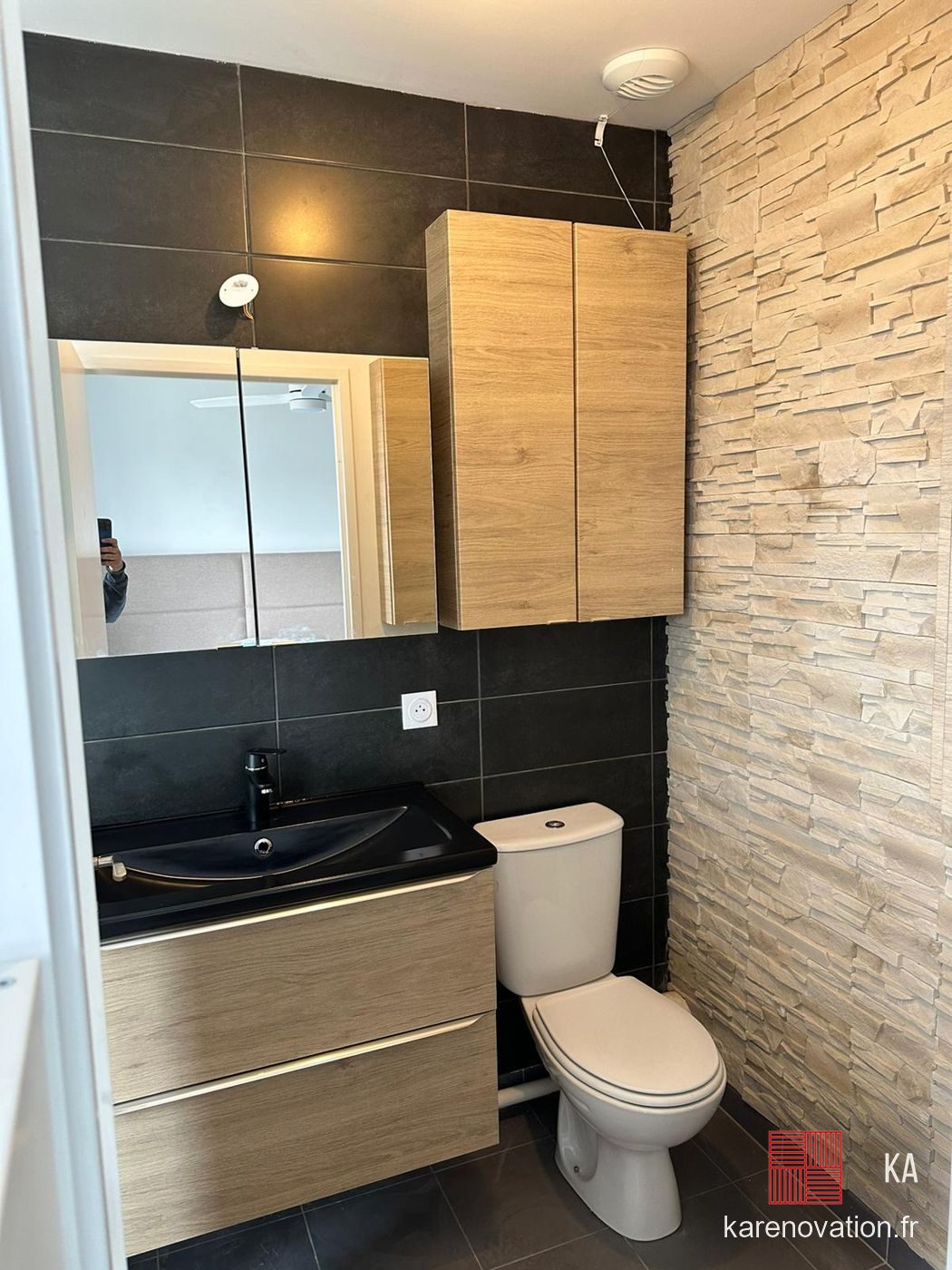 La même salle de bain après rénovation : mur effet pierre, vasque noire, meubles bois